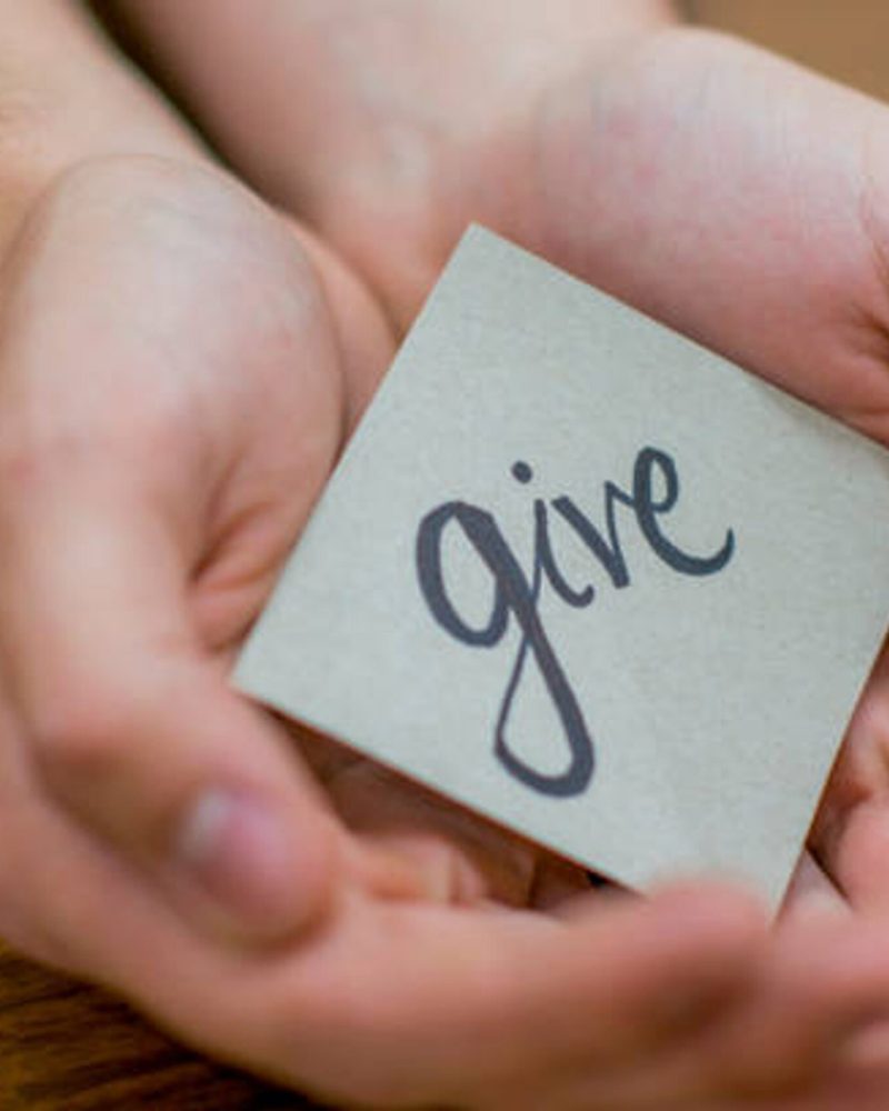 A man's hands rest on a wooden table and gently hold a small paper sign, which has the word 'give' written on it.